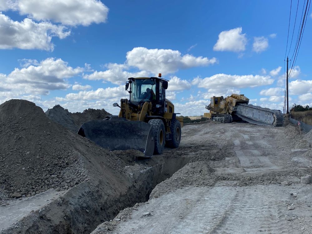 A loader pushes away a pile of newly unearthed rock and dirt removed by the large trencher in the background. The loader and trencher work in tandem during excavation activities for future wall construction near US 290 and Convict Hill Road. February 2022