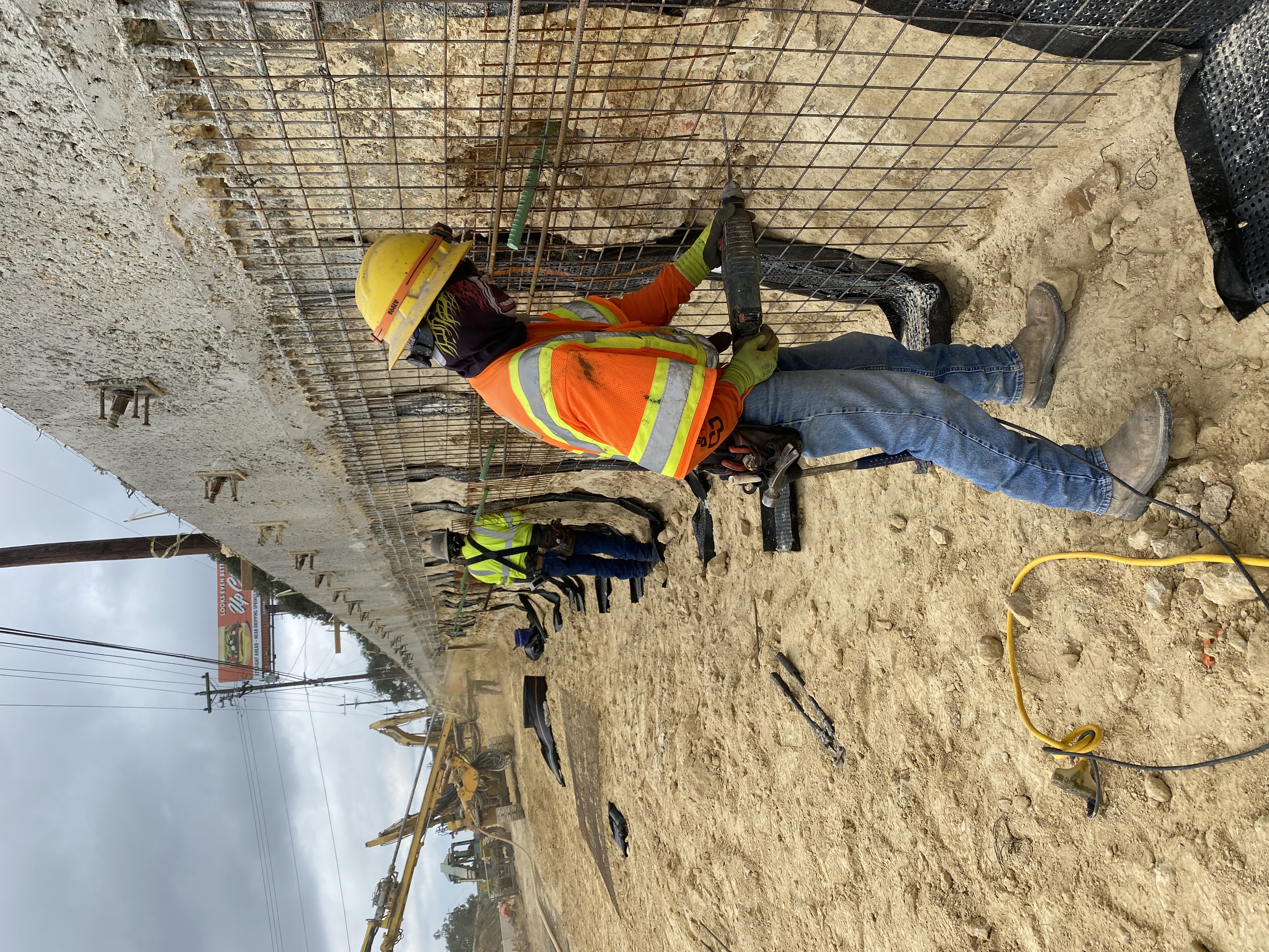 The team installs wire mesh reinforcements. Behind them a large soil nail drilling machine works to build a new retaining wall near US 290 and Circle Drive. December 2021