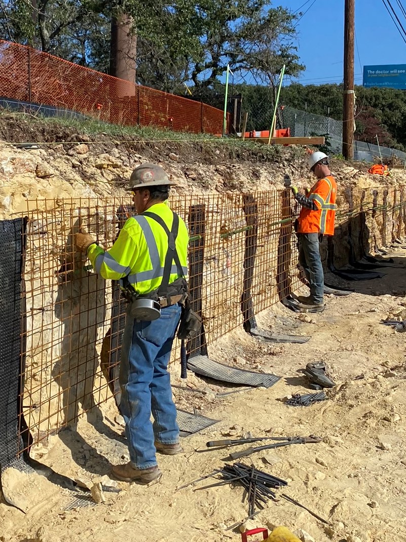 The team hangs wire mesh for a retaining wall on US 290 near Circle Drive. Above them, construction fencing has been placed as a safety measure and to separate work areas from nearby homes. December 2021