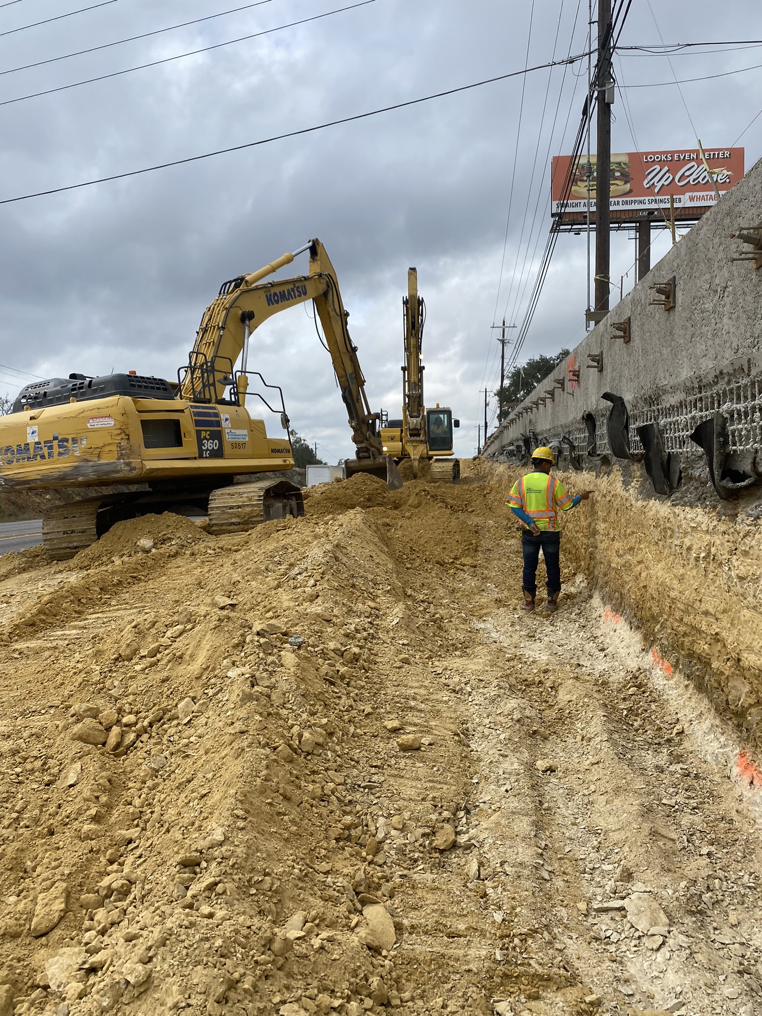 Excavators work to remove soil and rock next to completed sections of a retaining wall This retaining wall is built from top-to-bottom five feet at a time to hold rock in place while excavating to a lower grade. December 2021