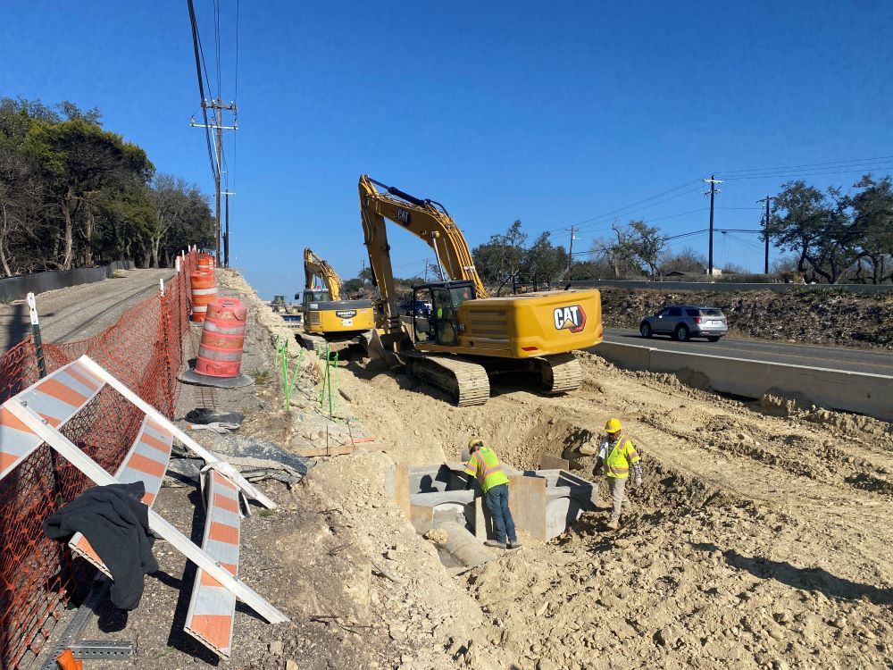 Oak Hill Parkway team members inspect recently placed drainage infrastructure as excavators work behind them to bury the rest of the underground pipe. This new drainage line sits underneath the location of a future westbound deceleration lane from US 290 